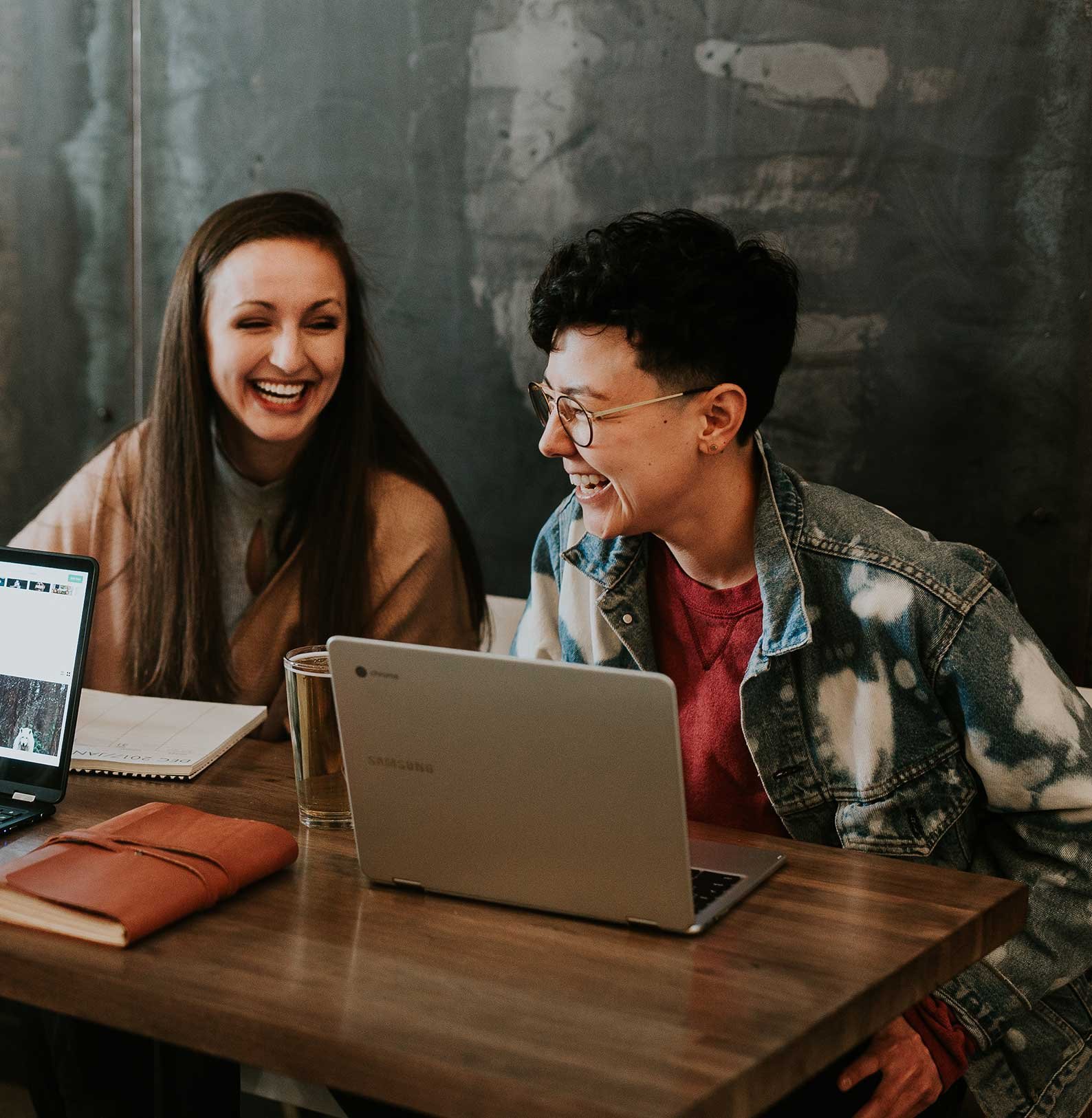 Two college students working on their computers in a coffee shop laughing.